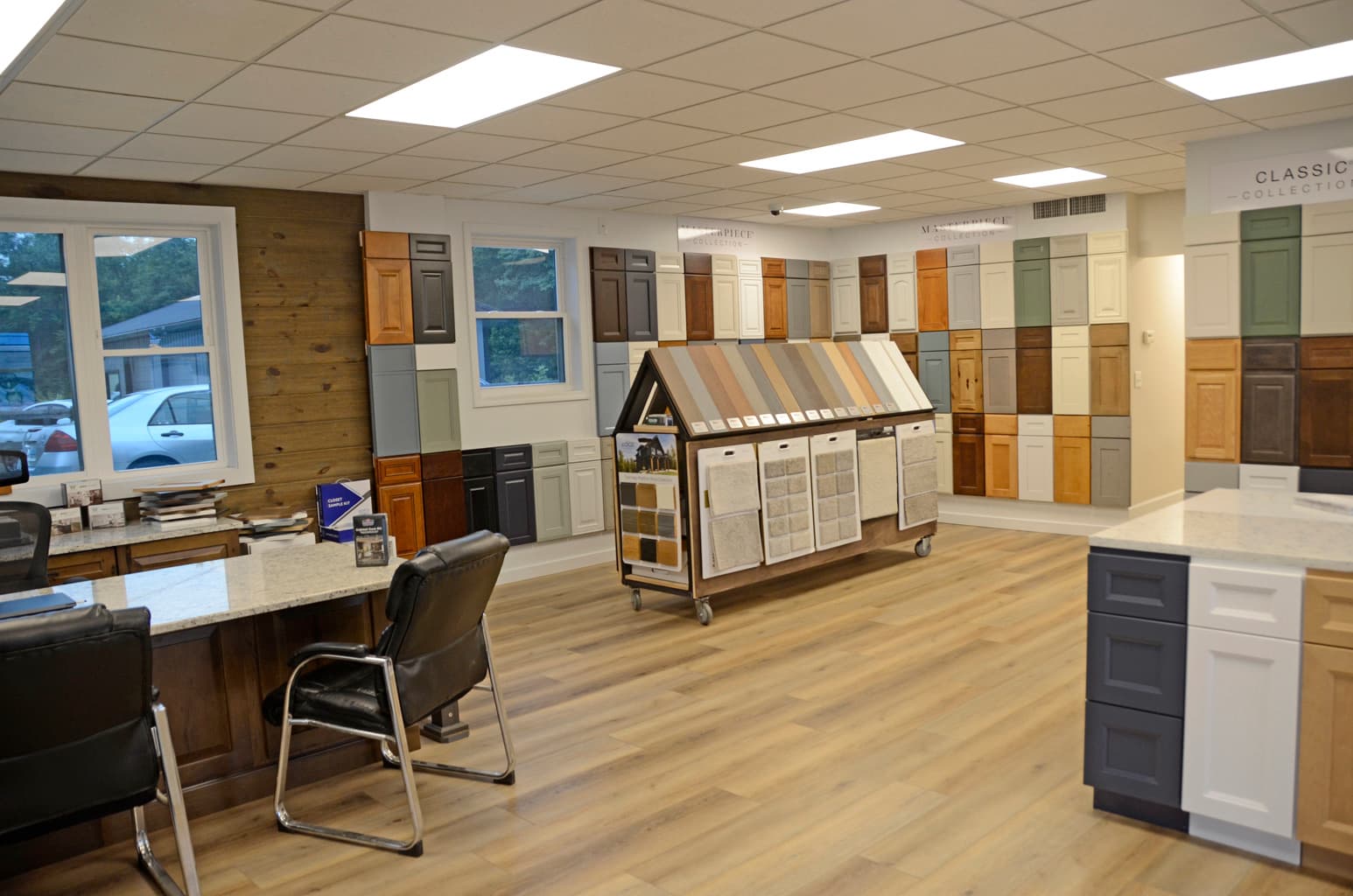 The Cabinets & More showroom floor, with cabinet door samples in white, grey, navy and stained oak lining the wall, a pull-out flooring sample display, and a granite-topped design desk.